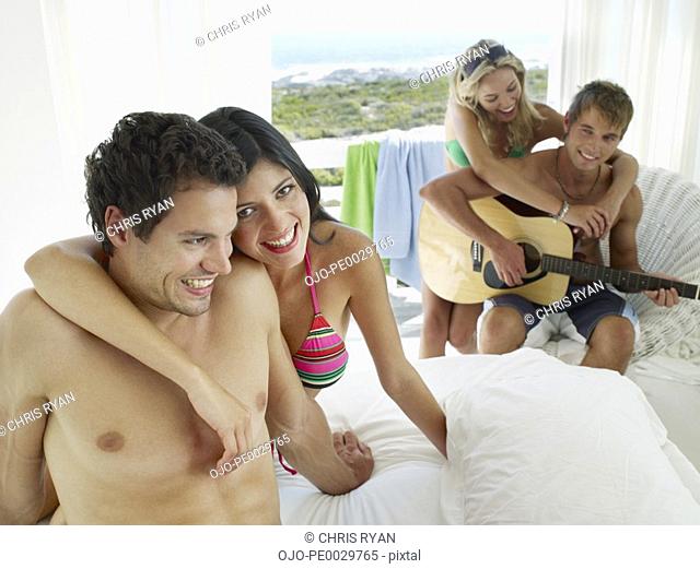 A couple sitting on a bed and another couple with an acoustic guitar