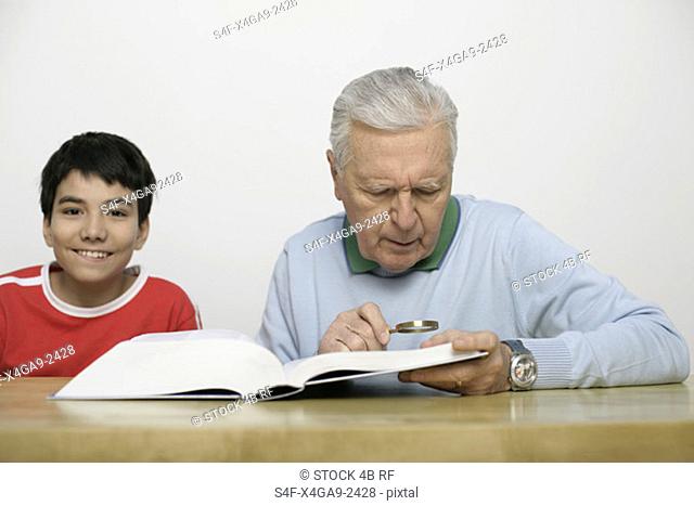 Grandfather using a magnifying glass to read a book, boy looking at camera, fully-released