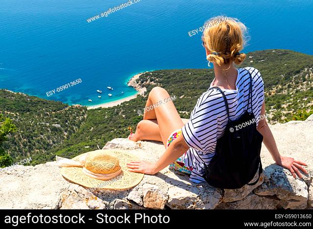 Active sporty woman on summer vacations sitting on old stone wall at Lubenice village, wearing straw hat and beach backpack enjoying beautiful coastal view of...