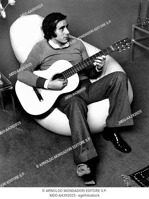 Roberto Vecchioni playing the guitar. Italian singer and songwriter Roberto Vecchioni sitting on a beanbag chair and playing the guitar. Milan, 1970s