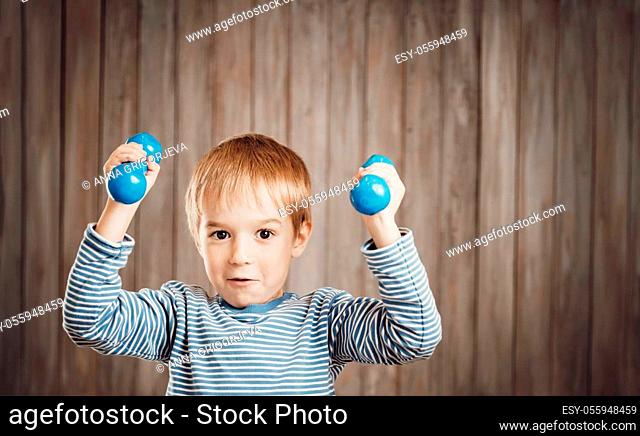 Child holding dumbbells indoors on wooden background. Happy active boy playing fitness. Healthy lifestyle