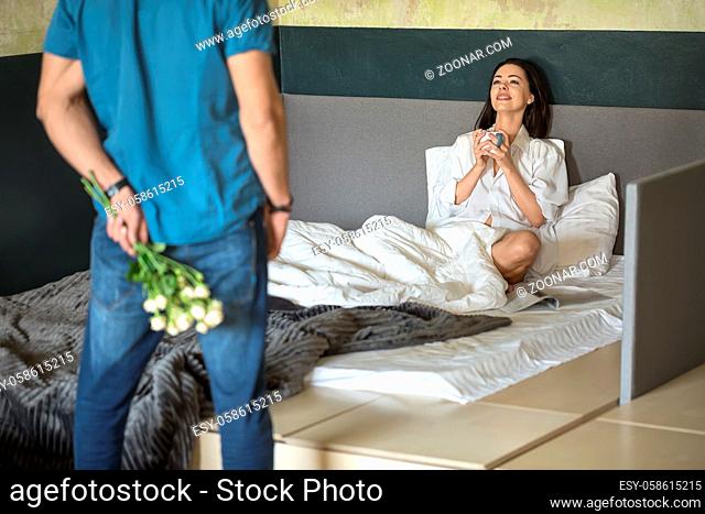 Happy smiling girl with a cup in the hands sits on the bed and looks at the man who holds a bouquet of flowers behind his back