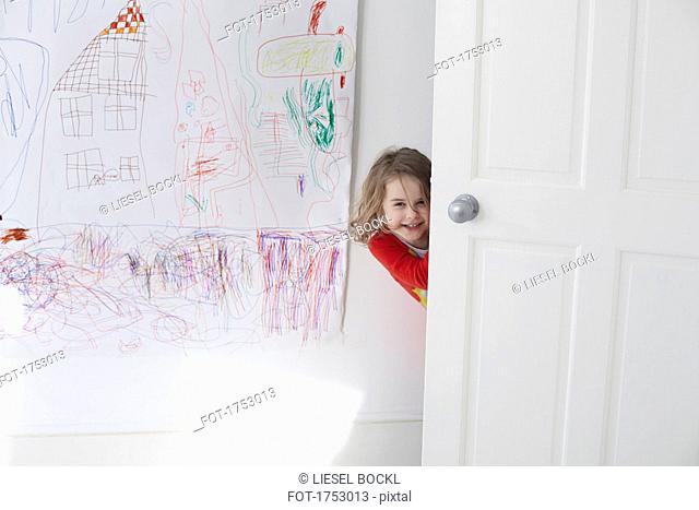 Portrait of cheerful girl hiding behind door against drawing on wall