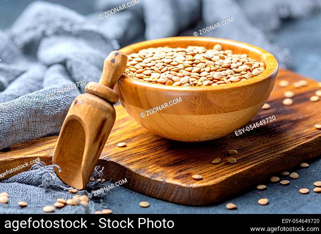 Bowl of brown lentils and a wooden scoop on a textured dark background, selective focus