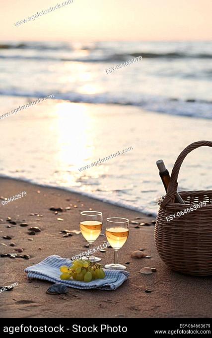 Two wine glasses with white wine standing on sand, on beach, beside grapes and wicker basket with bottle of wine