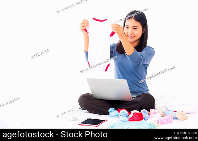 Asian mother shopping online for her baby clothes on laptop computer and tablet on the bed she makes purchase new baby clothes for an unborn baby preparing for...