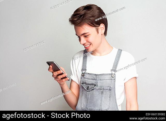 Portrait of happy handsome blogger teenager wearing in demin overalls and white T-shirt standing, using smartphone and texting message with toothy smile
