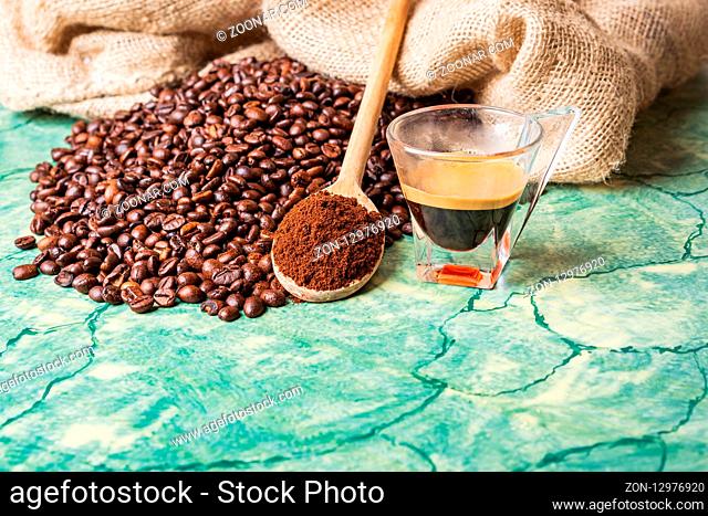 Coffee beans in coffee burlap bag on green surface, wooden spoon with ground coffee on top and coffee glass cup