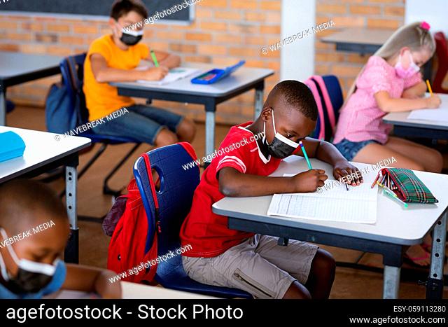 African american boy wearing face mask studying while sitting on his desk in the class at school