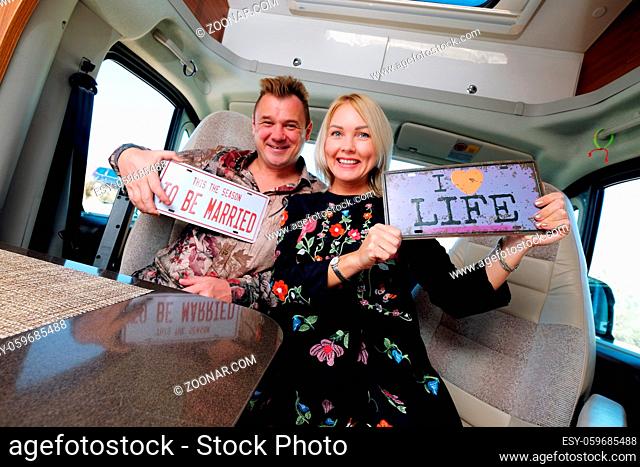 Cheerful laughing middle age family positive travelers couple sitting inside of camper holds hippy retro styled number plate or license number with text This...