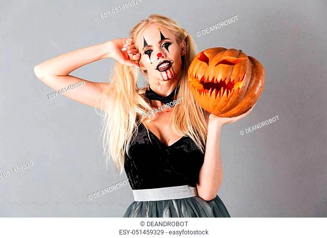 Young blonde woman in halloween make up posing with carved pumpkin and looking at the camera over gray background