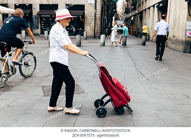 Senior woman walking on street while pushing shopping trolley bag in El Raval neighborhood, Barcelona, Catalonia, Spain