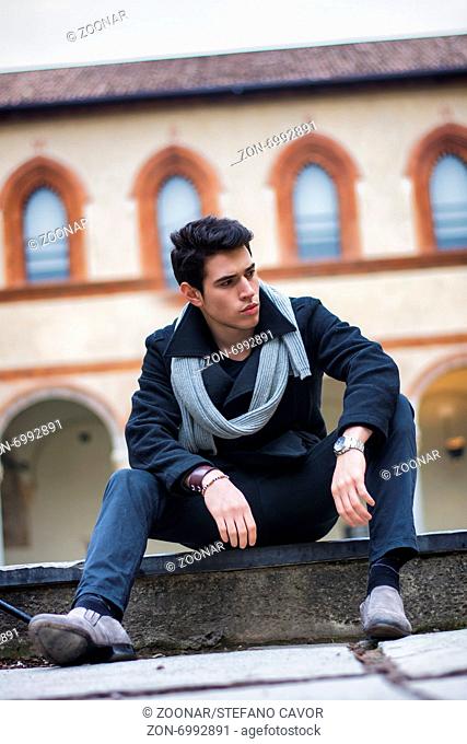 Stylish trendy young man sitting outdoor in old historical building looking to a side. Tilted shot