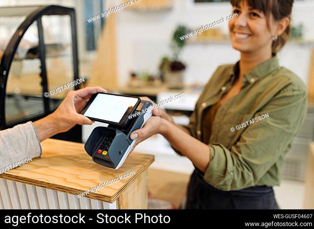 Cropped hand of customer doing contactless payment with smart phone to female owner at checkout in coffee shop