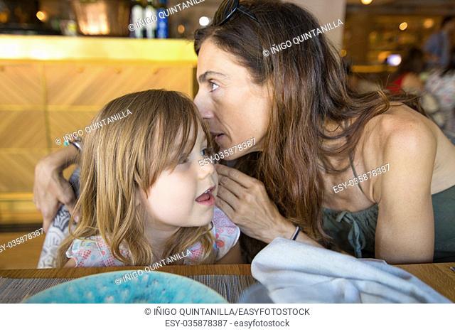 woman mother talking to ear to three years old blonde girl sitting in restaurant