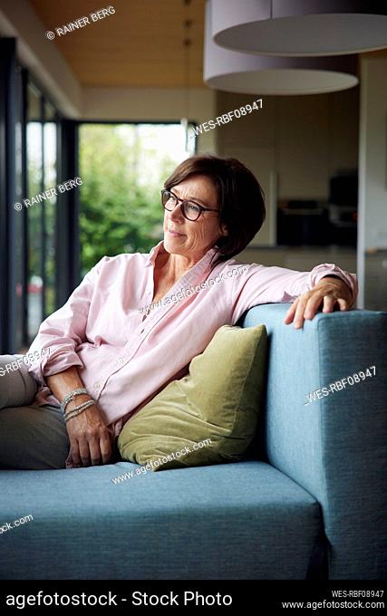 Thoughtful senior woman with eyeglasses sitting on sofa at home