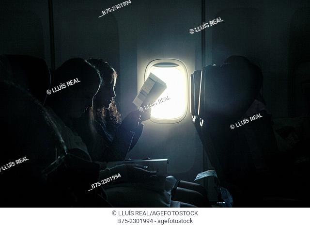 Two young girls sitting in the seats of an airplane reading a backlit of a window