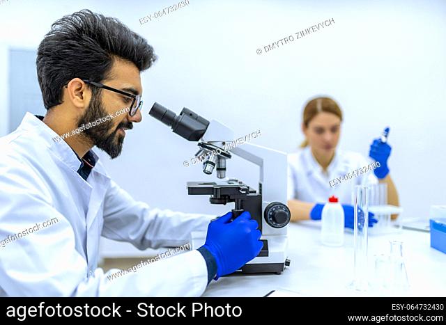 Health care researchers working in science laboratory, microscoping, looking focused at microscope, working together with colleague, making diagnostic