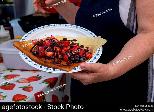 A closeup view of a person holding a dish with freshly prepared sweet crepes topped with strawberries, raspberries, blueberries and chocolate sauce