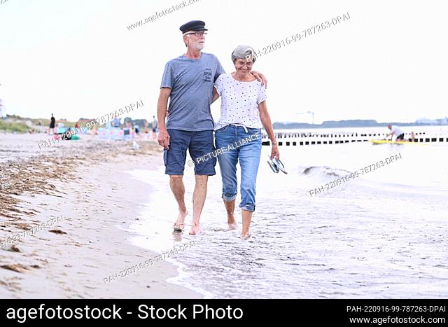 PRODUCTION - 20 August 2022, Mecklenburg-Western Pomerania, Rostock: ILLUSTRATION - A retired couple walks arm in arm on the beach