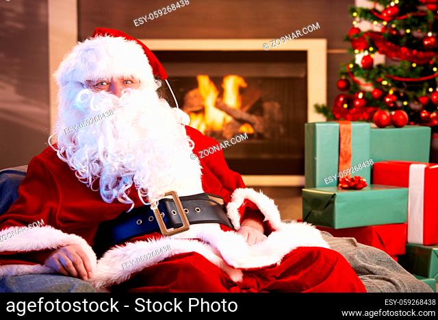 Portrait of Santa Claus sitting by fireplace with Christmas presents all around, looking at camera