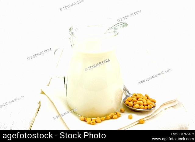 Soy milk in a jug, soybeans in a spoon on a napkin against the background of white wooden board