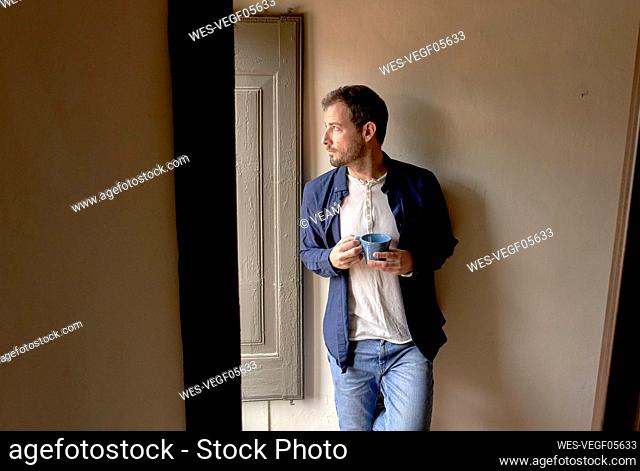 Thoughtful man with coffee cup leaning on wall looking out through window