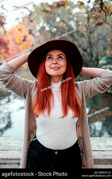 Smiling beautiful woman wearing hat looking up while standing against lake