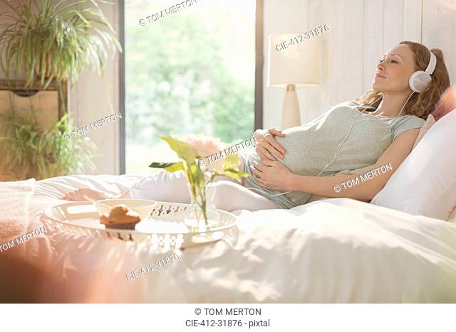 Serene pregnant woman listening to music with headphones on bed