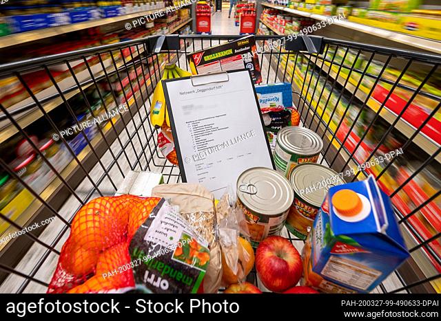 26 March 2020, Bavaria, Deggendorf: Food and a shopping list are in a shopping cart in a supermarket. Helpers provide food for the elderly and sick