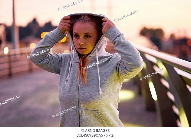 Curvaceous young woman training, getting hoody ready on footbridge at dusk