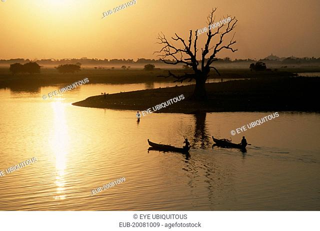 View from U Bein Bridge at Amarapura near Mandalay with canoes silhouetted against water reflecting golden sunset and trees and landscape in drifts of mist