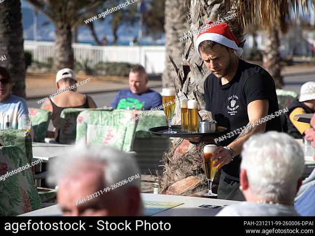 dpatop - 11 December 2023, Spain, Palma: Christian, a waiter at the restaurant ""Zur Krone"" on Arenal beach, wears a Santa hat during a sunny day with a record...