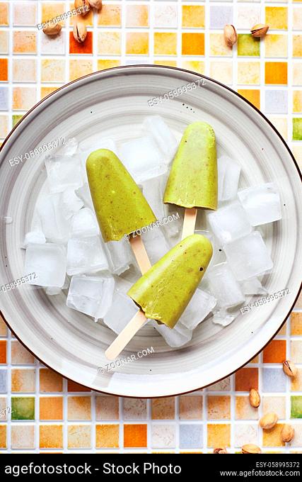 Frozen homemade pistachio popsicle in bowl of ice on mosaic tile table. Refreshing popsicle, frozen green juice on stick. Top view, copy space