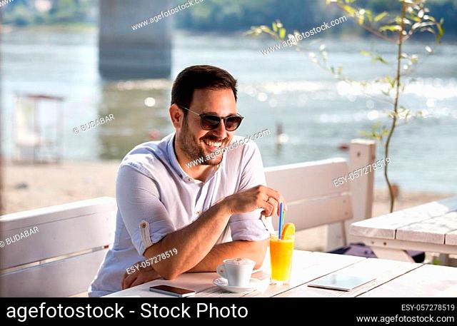 Handsome man with sunglasses drinking coffee and juice in cafe on beach. Enjoying morning beside river