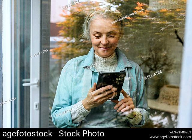 Smiling craftswoman looking at tablet PC in workshop seen through glass window