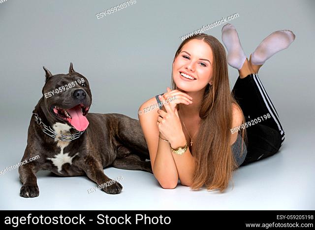 Beautiful sporty young woman lying on floor with adult grey amstafford terrier dog. Studio shot over gray background. Copy space