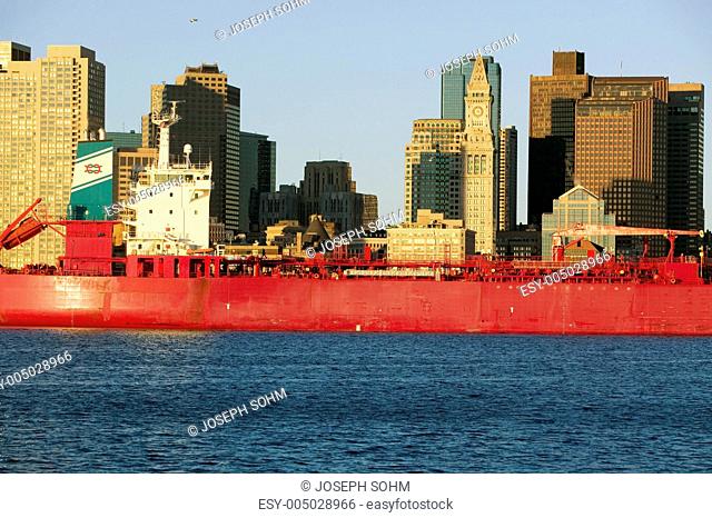 Bright red cargo ship travels in front of Boston Harbor and the Boston skyline at sunrise as seen from South Boston, Massachusetts, New England