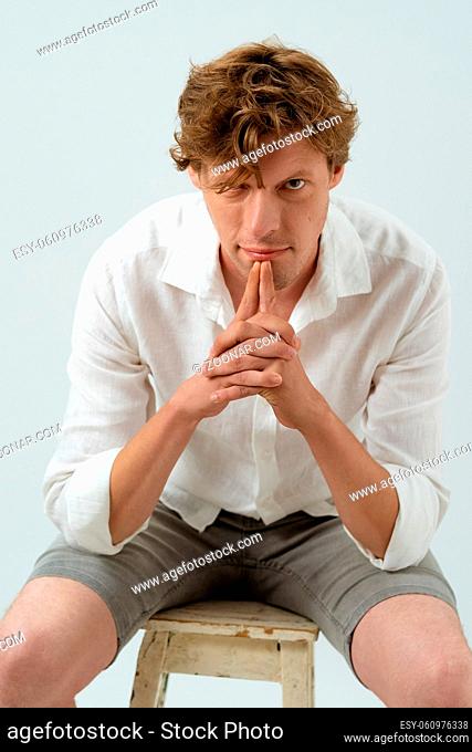 Confident young handsome man sitting on chair pensive looking at camera with hands folded and fingers at the chin isolated on white background