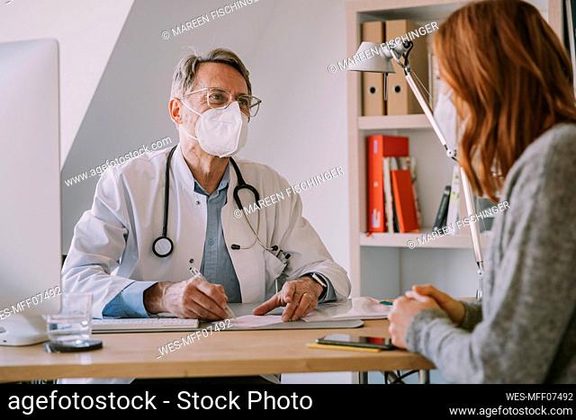 Male general practitioner with face mask looking at patient while sitting at doctor's office