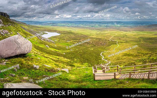 Large panorama with view on steep stairs of wooden boardwalk leading to Cuilcagh Mountain peak with lake and valley or plane below, Northern Ireland