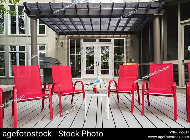 Puertas de patio y estructura de pérgola marrón sobre terraza de madera con sillones de lona roja en la parte trasera de la casa revestida de tablones de madera...