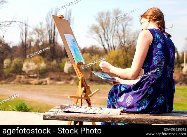 Young woman creative painter draws brush watercolor paints on canvas outdoors. Spring inspiration. Female artist painting landscape