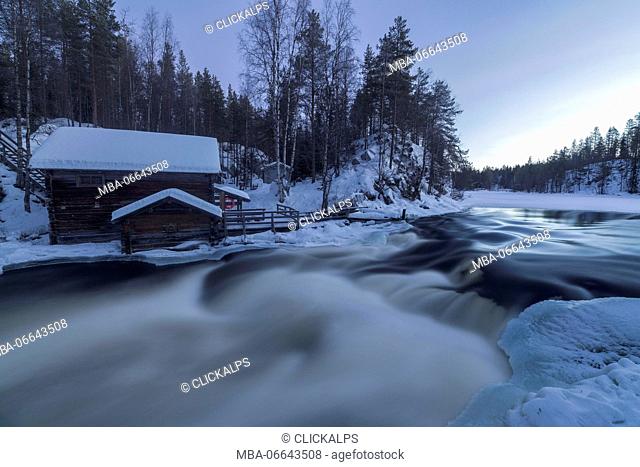 A wooden hut surrounded by the river rapids and snowy woods at dusk Juuma Myllykoski Lapland region Finland Europe