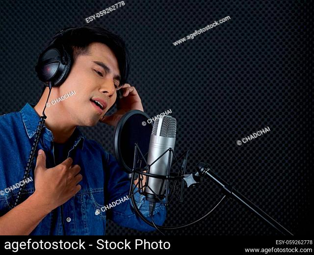 Young asian man with headphone singing in front of black soundproofing wall. Musician producing music in professional recording studio