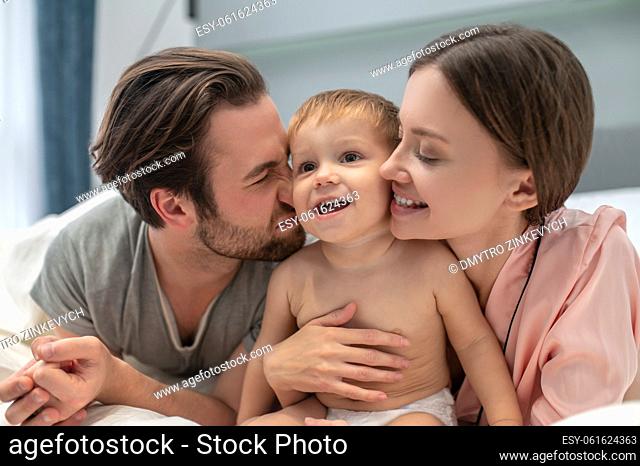 Happiness. Young adult happy man and woman with closed eyes touching cute baby looking up on bed in bedroom