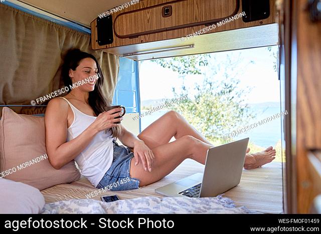 Smiling woman with coffee cup watching laptop in camper van