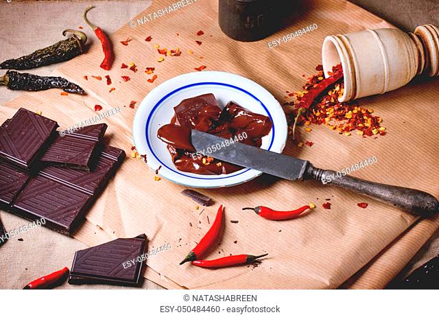 Chopping dark chocolate with fresh and dry red hot chili peppers on baking paper with plate of hot melted chocolate and vintage knife