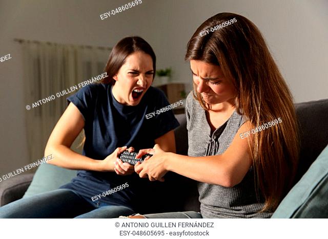 Two friends fighting for the tv remote control sitting on a couch in the living room at home