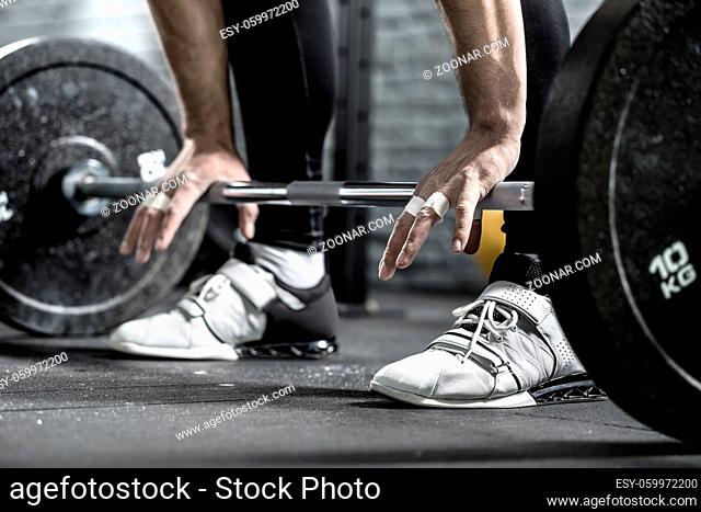 Barbell on the floor and the man's hands on it on the background of guy's legs in the black pants and white sneakers. Close-up horizontal photo